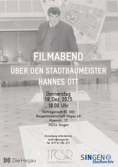 Ein Mann in dunklem Anzug steht hinter einem Rednerpult in einem Sitzungssaal; im Hintergrund sitzen weitere Personen an einer langen Tafel, während auf einer großen Wandkarte ein Poster mit dem Titel Filmband über den Stadtbaumeister Hannes Ott und Veranstaltungsangaben zu sehen ist.